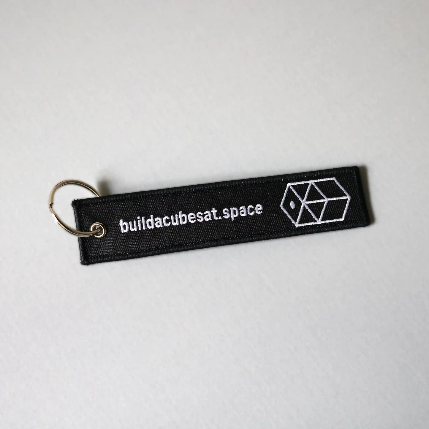 Black keychain with white text and logo on the front on a light gray background, slightly tilted to the left