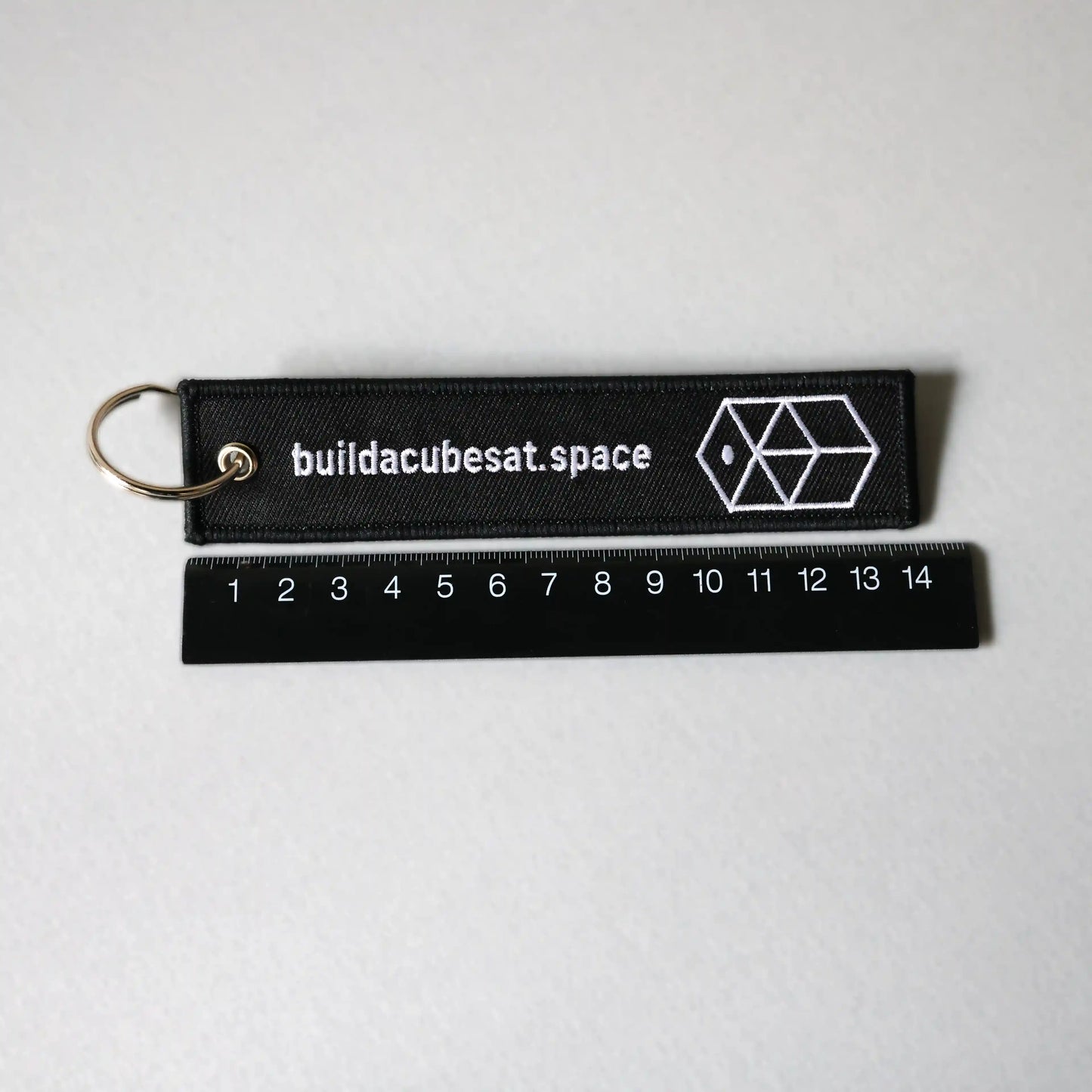 Black keychain with white text and logo on a gray background and a black ruler for scale indication