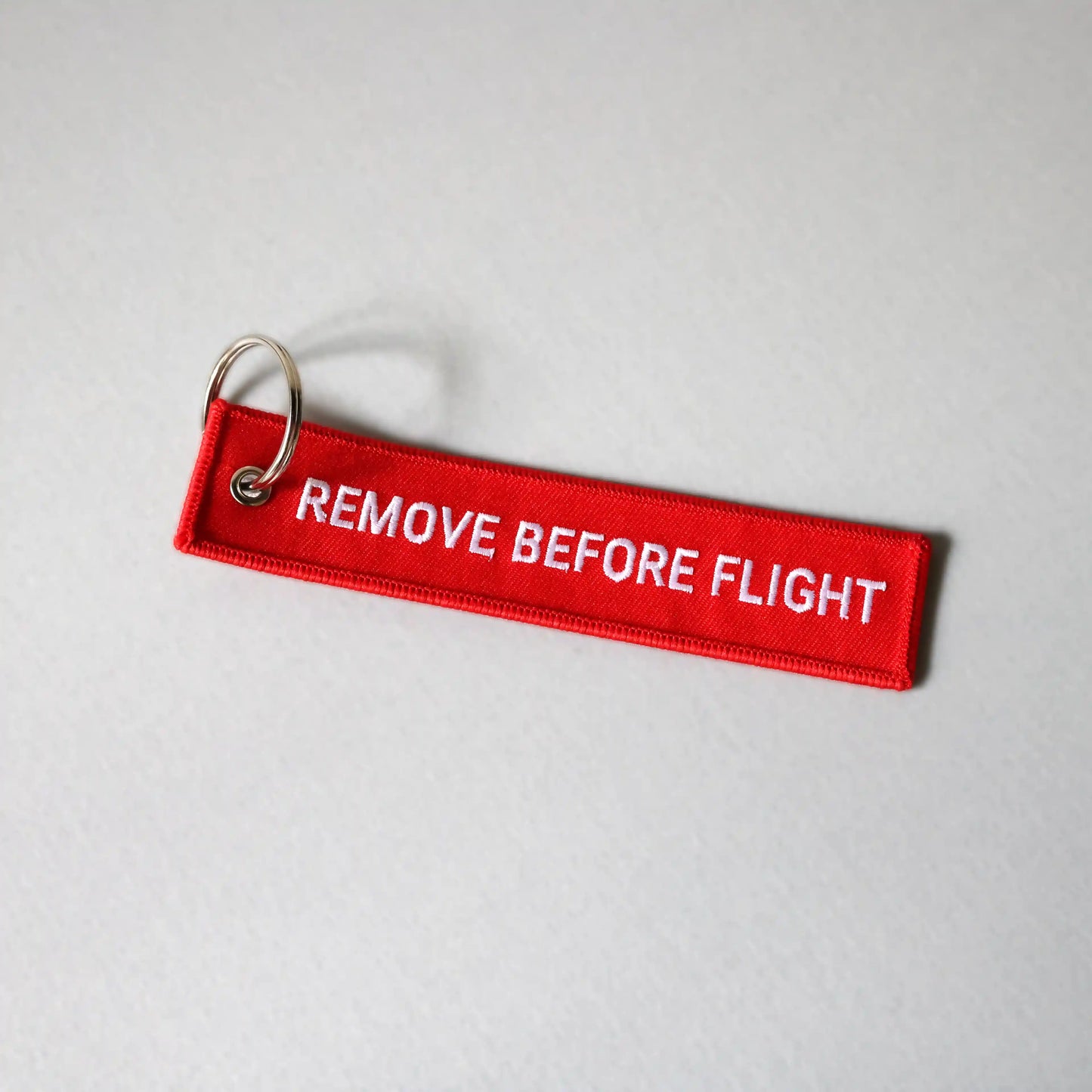 Red 'REMOVE BEFORE FLIGHT' keychain on a gray background, tilted to the right
