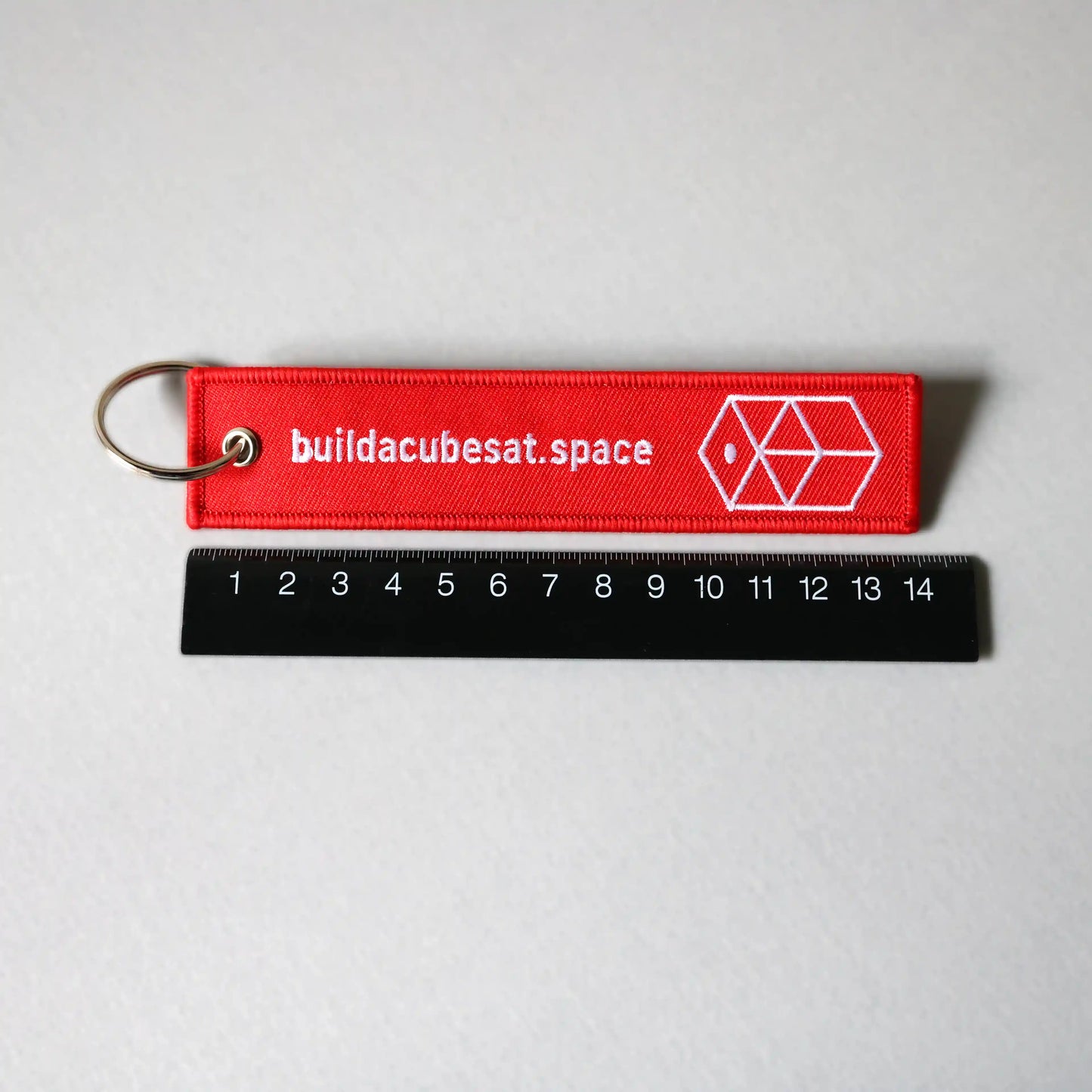 Red keychain with text and logo on a gray background, with a ruler for scale indication