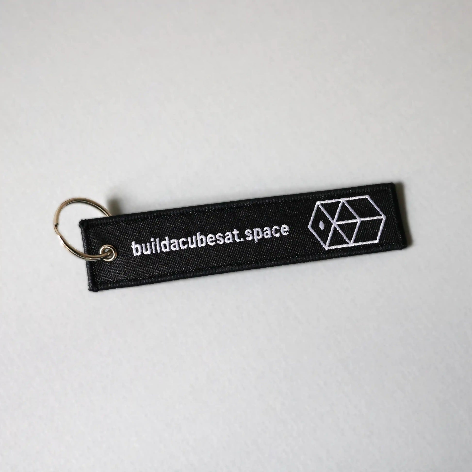 Black keychain with white text and logo on the front on a light gray background, slightly tilted to the left