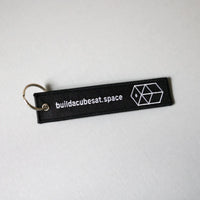 Black keychain with white text and logo on the front on a light gray background, slightly tilted to the left