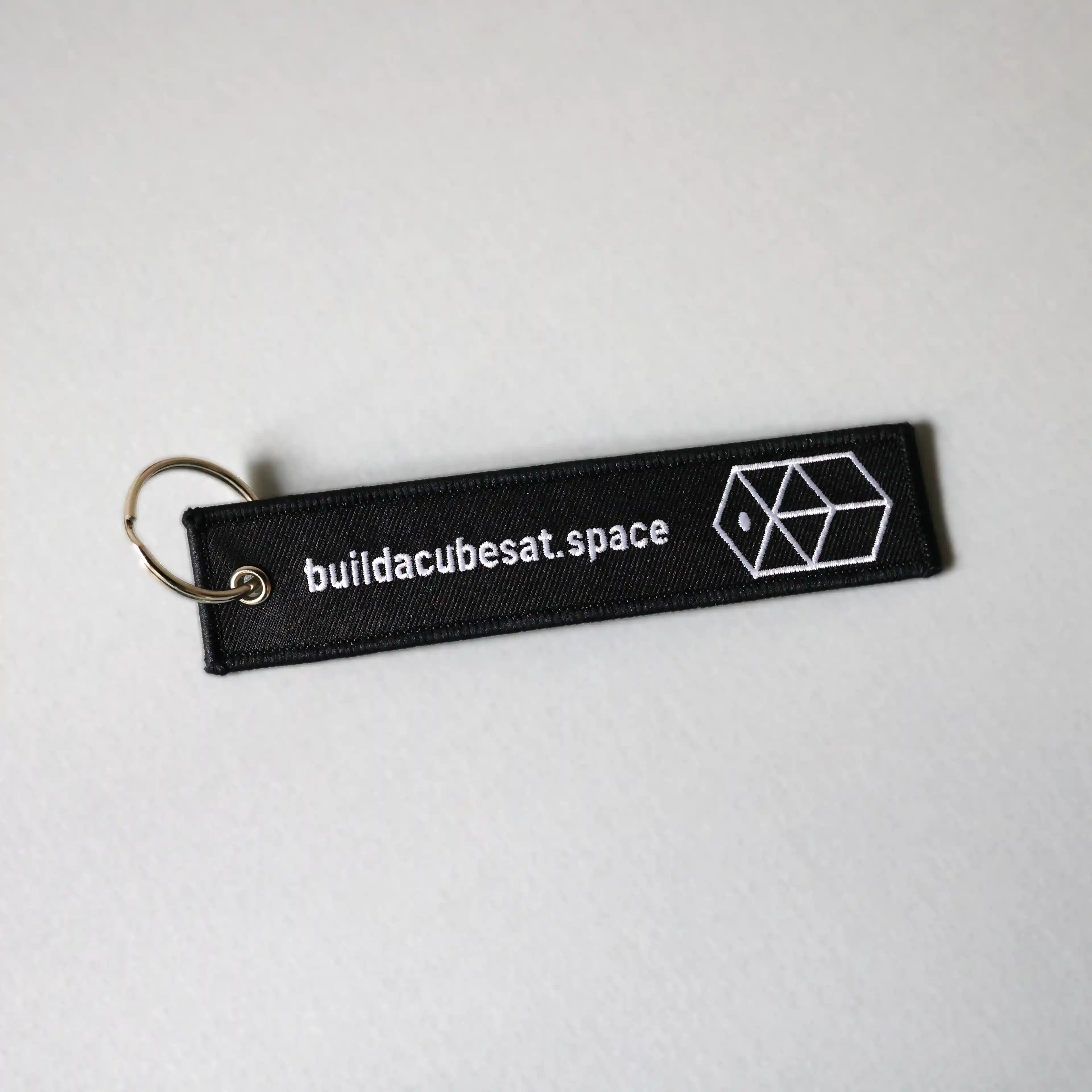 Black keychain with white text and logo on the front on a light gray background, slightly tilted to the left
