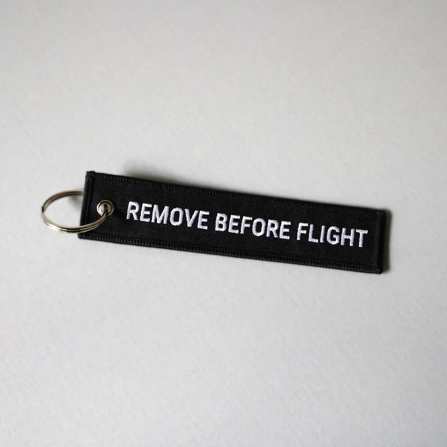 Back side of a black keychain with 'REMOVE BEFORE FLIGHT' text on a light gray background, tilted to the right