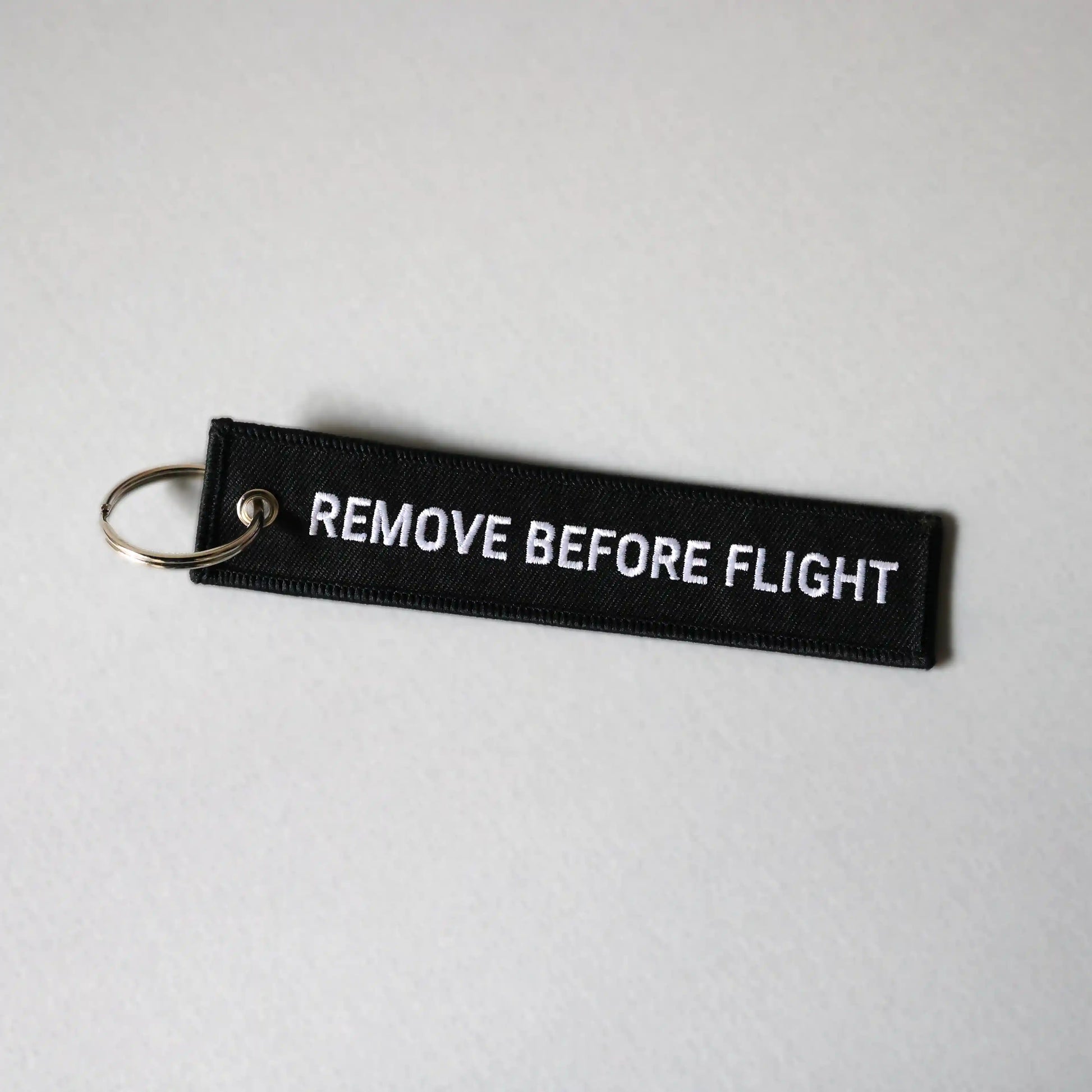 Back side of a black keychain with 'REMOVE BEFORE FLIGHT' text on a light gray background, tilted to the right
