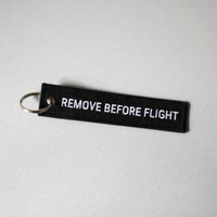 Back side of a black keychain with 'REMOVE BEFORE FLIGHT' text on a light gray background, tilted to the right