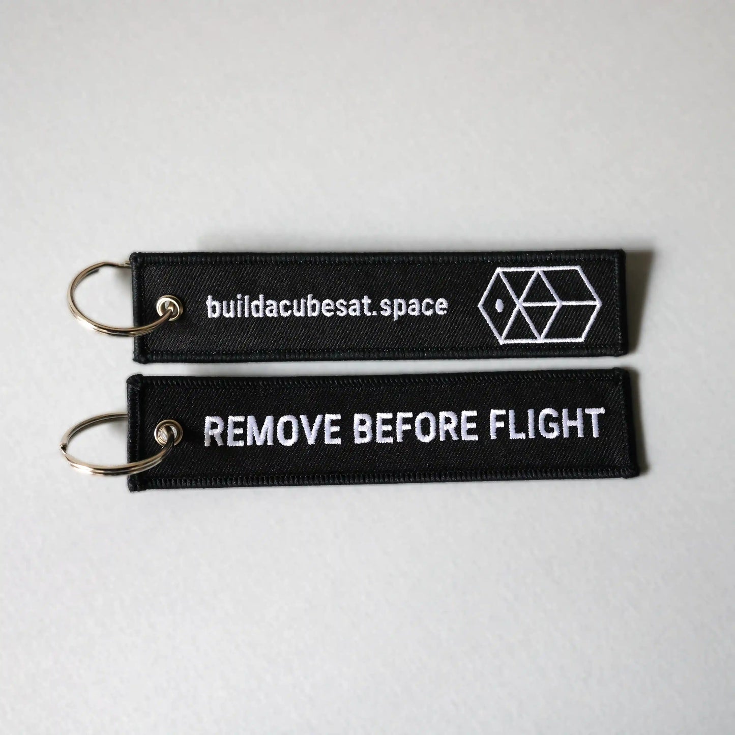 Two black keychains with white text on a gray background, showing the front and back sides of the product