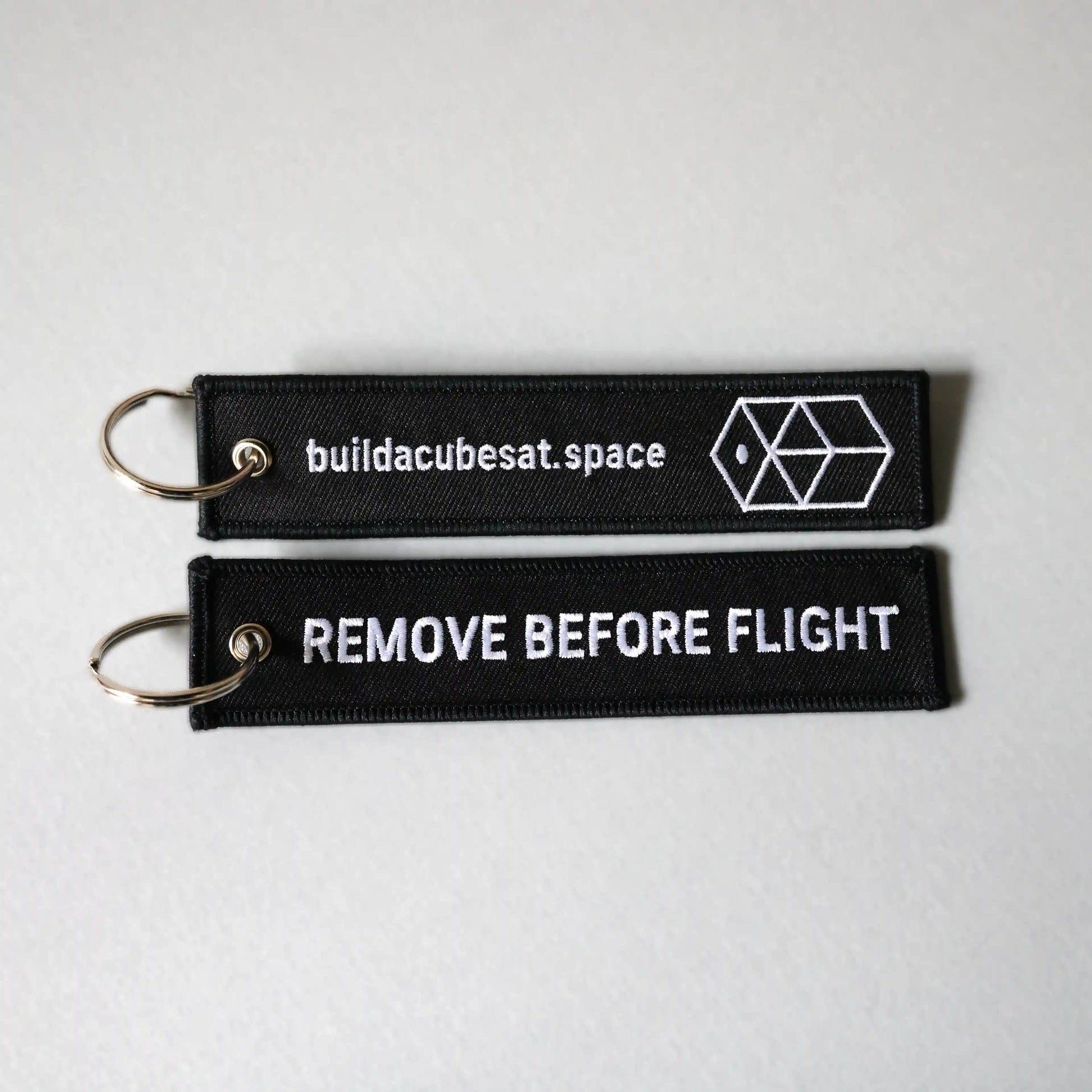 Two black keychains with white text on a gray background, showing the front and back sides of the product