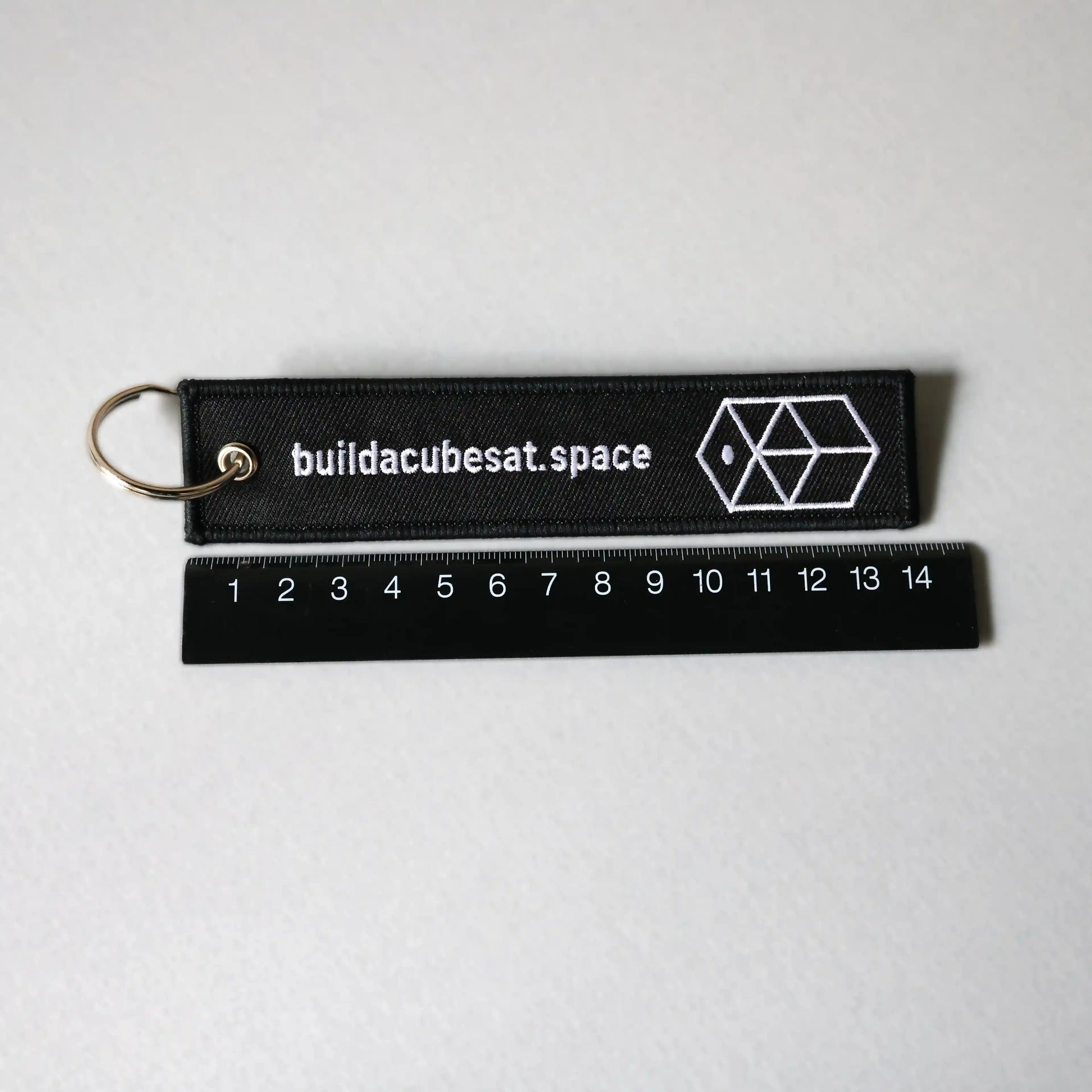Black keychain with white text and logo on a gray background and a black ruler for scale indication