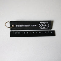 Black keychain with white text and logo on a gray background and a black ruler for scale indication