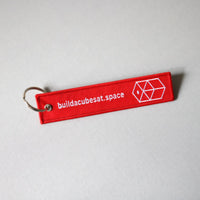 Red keychain with white text and logo on the front side on a light gray background, tilted to the left