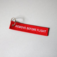 Red 'REMOVE BEFORE FLIGHT' keychain on a gray background, tilted to the right